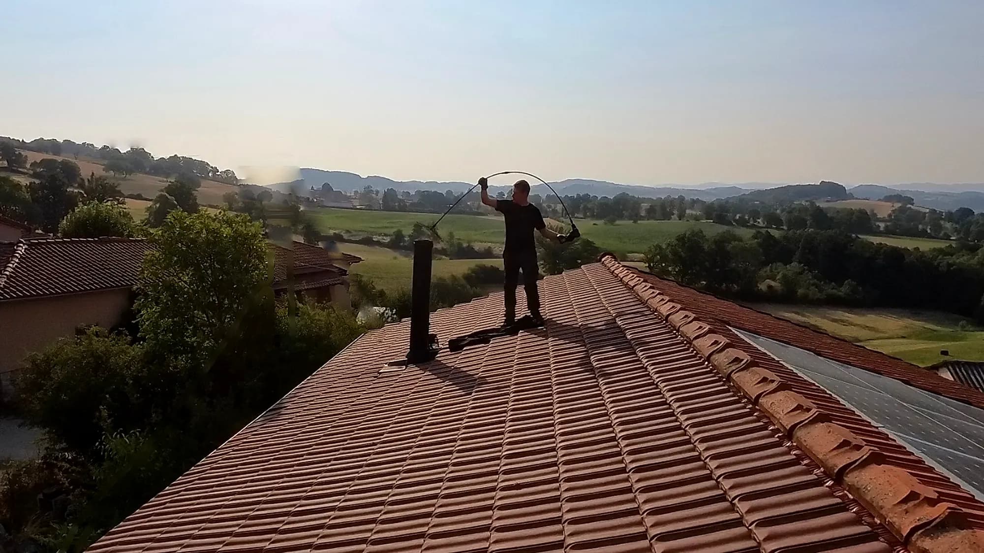 Ramoneur de Ramoneurs de France en intervention sur le toit d'une maison, canne rotative en main, au-dessus de la campagne de la Loire